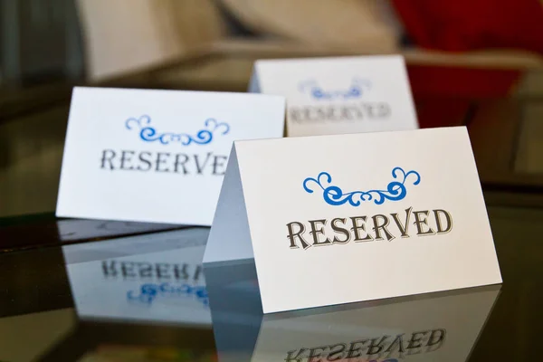 Reserved signs on reflective glass surface, creating depth effect. Elegant serif RESERVED sign in focus with ornate blue swirl designs. Softly lit indoor setting suggests exclusivity. Ideal for