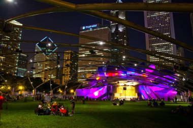 Chicago, Illinois 'de akşam karanlığında canlı bir şehir parkı, mor ve sarı ışıklarla parlayan modern bir amfi tiyatronun bulunduğu, çeşitli bir topluluk tarafından çevrelenmiş, açık havada sakin bir akşamın keyfini çıkaran,