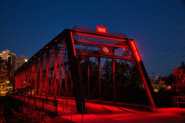 Çarpıcı kırmızı ışıklı tarihi kiriş köprüsü alacakaranlıkta, karmaşık metal kalıpları ve geometrik kalıpları sergiliyor, kentsel bir ufuk çizgisine karşı kurulmuş.