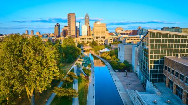 Indianapolis 'te Altın Saat Parlıyor - Günbatımında canlı şehir manzarasını modern mimari ve yemyeşil ile kesen sakin şehir kanalının havadan görünüşü