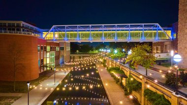 2023 'te Indianapolis' teki modern aydınlık yaya köprüsü yansıtıcı kanal ve canlı kampüs yolunun akşam görüntüsü.
