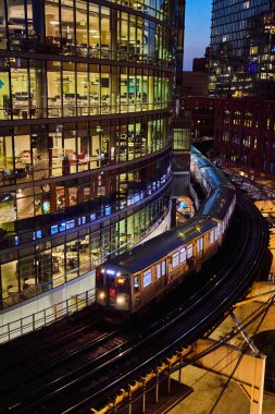 2023 'te Chicago' nun Alacakaranlık Şehir Manzarası 'nda yükselen raylarda parlayan bir tren ve şehir ışıklarını yansıtan modern bir ofis binası yer alıyor.