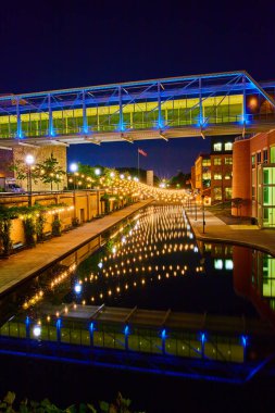 2023 yılındaki canlı Indianapolis gece manzarası, sakin kanal suları üzerinde aydınlık bir yaya köprüsü sergiliyor, canlı gezinti alanları ve karışık kullanım amaçlı binalarla çevrili..