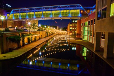 Canlı Indianapolis gece manzarası yansıtıcı kanal üzerinde mavi ışıklı bir yaya köprüsü sergiliyor, aydınlık modern binalarla çevrili, 2023