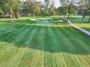 Fort Wayne, Indiana 'da titizlikle sürdürülen sakin bir golf sahasının havadan görünüşü canlı yeşil çim, kum sığınakları ve yumuşak sabah güneşi altında dönen bir golf arabası yolu..
