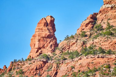 Berrak mavi gökyüzünün altında canlı kırmızı kumtaşı kulesi, Sedona, Arizona 'da yeşilliklerle çevrili, Güneybatı güzelliğinin simgesi.