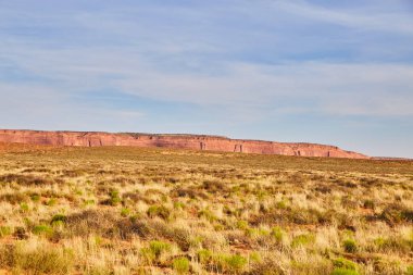 Anıt Vadisi, Arizona, 2016 'da sakin bir öğleden sonra. Geniş bir çalı tarlası ve bulutlu bir gökyüzü altında heybetli kırmızı bir mesa..