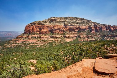 Devils Köprüsü, Sedona, Arizona 'daki görkemli kırmızı kaya oluşumlarının nefes kesici görüntüsü, açık mavi bir gökyüzü altında, yemyeşil, doğal güzellikleri ve jeolojik harikaları vurguluyor..