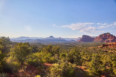 Sedona, Arizona 'daki Bell Rock' ın nefes kesici manzarası katmanlı kırmızı kaya oluşumları, yemyeşil yerel bitki örtüsü ve öğleden sonra aydınlatması. Doğa, macera ve seyahat temaları için mükemmel.