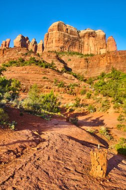 Sedona, Arizona 'daki Katedral Kayası' nın gündüz manzarasında canlı kırmızı kaya oluşumları, bir cairn ile işaretlenmiş popüler bir yürüyüş yolu ve seyrek çöl bitkileri yer almaktadır..