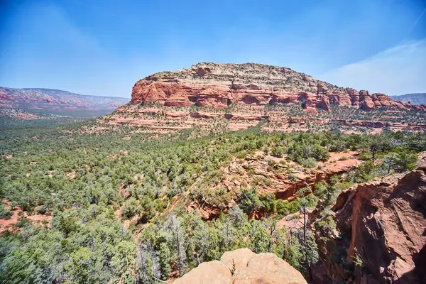 Sedona 'daki Arizonas Devils Köprüsü' nün nefes kesici gündüz görüntüsü, açık mavi gökyüzü altında yükselen kızıl kaya oluşumlarının zemininde yeşillik sergiliyor.