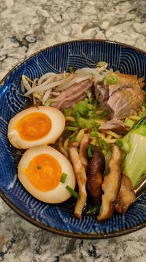 Canlı, leziz domuz eti ve taze sebzeli ramen kasesinin göz hizasında yakın plan görüntüsü mermer bir tezgahta sanatsal olarak sunuluyor..