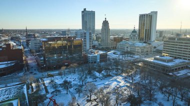 Fort Wayne, Indiana 'da kış sabahı, çeşitli bir şehrin silueti arasında kar örtülü bir park sergileniyor.