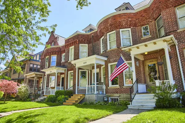 stock image Sunny suburban street in Fort Wayne with traditional brick houses and American flag, symbolizing community and patriotism.