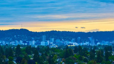 Portland, Oregon şehir merkezinin altın saatinde havadan görünüşü modern yüksek binaların ve yemyeşil yeşilliklerin sergilendiği sakin bir gökyüzü altında kayan tepeler ve yoğun bir orman arkaplanı..