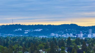 Portland 'da sabah dinginliği: Şehir merkezinin panoramik hava manzarası, modern yüksek binalar ve pastel gündoğumu renkleriyle yıkanmış, verimli bir orman yamacına karşıdır. İdeal