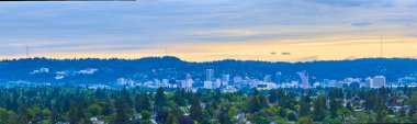 Portland, Oregon şehir merkezinin havadan görünüşü, modern gökdelenlerin ve yemyeşil yeşilliklerin bir karışımını ormanlık tepelerin arka planına karşı sergiliyor, kentsel ve doğal manzaraların dingin güzelliğini yakalıyor.