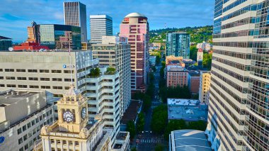 Portland, Oregon şehir merkezinin hava manzarası ofis binaları, gökdelenler ve tarihi saat kulesiyle dolu canlı bir şehir manzarası sergiliyor. Modern mimarinin ve yemyeşil tepelerin karışımı