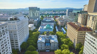 Portland şehir merkezinin havadan görünüşü, tarihi adliye binasının çevresi yemyeşil ağaçlar ve modern gökdelenlerle çevrili. Manzaralı arka planda akan bir nehir ve uzak dağlar yer alıyor. İdeal