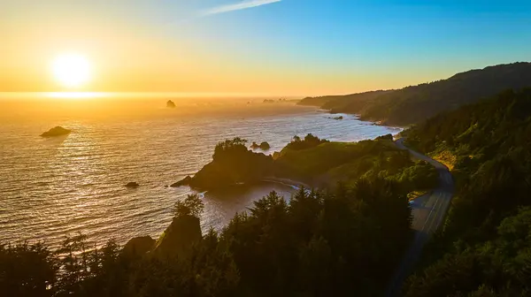 stock image Aerial view of Arch Rock in Brookings, Oregon during sunset. The golden hour light highlights the meandering coastal road, dramatic cliffs, and tranquil ocean in the Samuel H. Boardman State Scenic