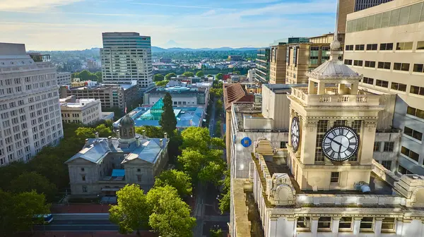 stock image Aerial view of Portlands downtown, featuring the historic clock tower of the Pioneer Courthouse and the neoclassical dome surrounded by lush greenery, with modern high-rises and scenic mountains in