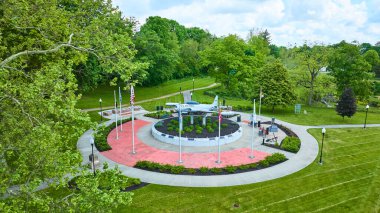 Aerial view of Huntington Indianas Memorial Park showcasing a vintage U.S. Air Force aircraft display, lush green trees, and symmetrical flag arrangements. A serene tribute to history and nature. clipart