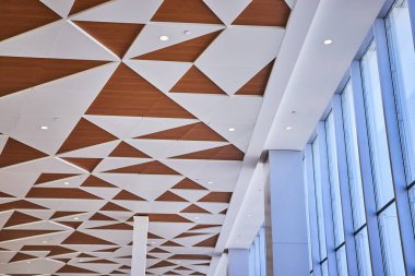 Beyaz ve ahşap tonlu üçgenlerden oluşan geometrik bir tavanı olan modern havaalanı içi. Yüksek pencerelerden geçen doğal ışık, mimari zarafeti ve yenilikçi tasarımı vurgular.