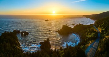 Brookings, Oregon 'daki Arch Rock' ın hava görüntüsü, gün batımında dolambaçlı kıyı yolu ve yoğun yeşil ormanları yakalıyor. Altın saat ışığı Samuel H 'in sükunetini ve doğal güzelliğini arttırır.