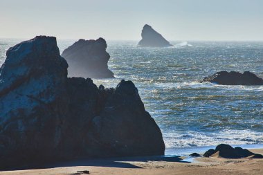 Serene coastal scene at Harris Beach State Park, Oregon. Rugged dark rocks dominate the foreground while the shimmering blue ocean and white waves in the background evoke a sense of escape and clipart