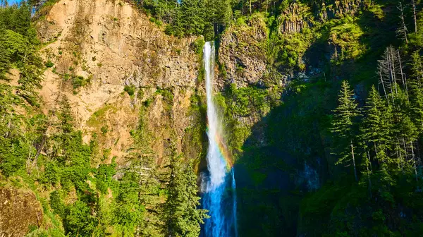 stock image Majestic Multnomah Falls in Oregons Columbia Gorge cascades into a serene pool, surrounded by lush forests and rugged cliffs. A striking rainbow arcs gracefully through the mist, creating an ethereal