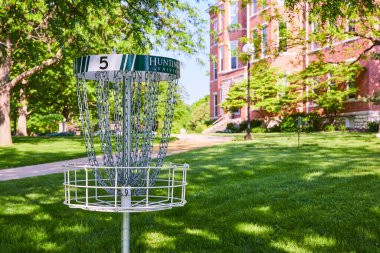 Serene Huntington Üniversitesi kampüs sahnesinde yemyeşil çimlerin üzerinde bir disk golf sepeti, olgun ağaçlarla çevrili ve arka planda kırmızı tuğlalı bir bina. Eğitim temaları ve öğrenim hayatı için ideal