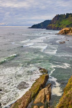 Oregon, Devils Punchbowl 'da sakin deniz manzarası. Bereketli yeşilliği ile engebeli uçurumlar çalkantılı mavi-yeşil denize iniyor. Dalgalar dinamik bir gökyüzü altında sivri kayalara karşı kırılır ve huzur yaratır.
