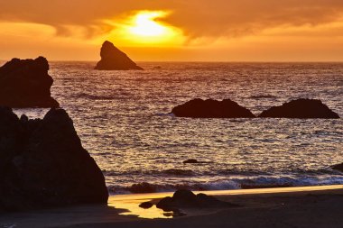 Brookings, Oregon 'daki Harris Beach Eyalet Parkı' nda altın gün batımı. Güneş, engebeli kaya oluşumlarına ve sakin okyanuslara sıcak bir parıltı yayar. Huzurlu bir kıyı manzarası oluşturur.