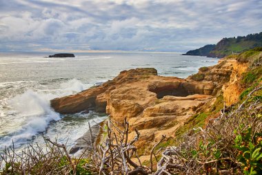 Canlı turuncu ve kahverengi tonlara sahip engebeli sahil kayalıkları Otter Rock, Oregon 'daki Devils Punchbowl' da köpüklü dalgalarla buluşuyor. Fırtınalı bir gökyüzü ve uzak bir ada Bu nefes kesici derinliğe dram ve derinlik katar