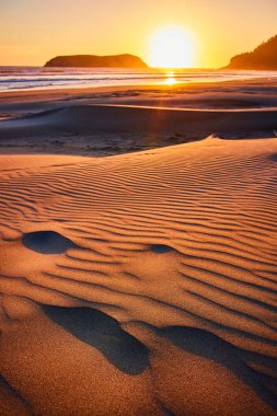 Gold Beach, Oregon 'da günbatımı, dalgalı kum desenleri ve nazik okyanus dalgalarıyla altın renkleri ile sakin bir sahne çizer. Silüetli bir kaya oluşumu derinlik katar. Huzurlu ve hayranlık uyandırıcı bir şey yaratır.