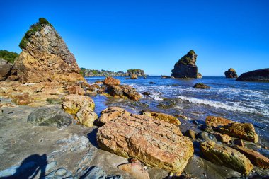 Oregon 'daki Harris Beach Eyalet Parkı' nda nefes kesici sahil manzarası. Ön planda engebeli kayalar, karada heybetli deniz yığınları ve parlak mavi gökyüzü çarpıcı bir görüntü yaratır. Seyahat için ideal, doğa.