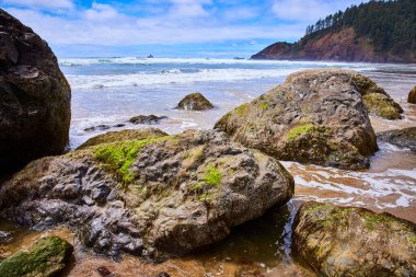 Indian Beach, Oregon 'daki dinamik kıyı sahnesi: yosun kaplı kayalar parçalı bulutlu bir gökyüzünün altında okyanus dalgalarıyla çarpışıyor. Seyahat, çevre ya da eğitim için mükemmel. Tecrübe doğası