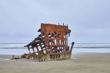 Rusted remnants of the Peter Iredale shipwreck on a sandy beach in Hammond, Oregon. Under overcast skies, the corroded iron beams stand against the ocean waves, a silent testament to maritime history clipart