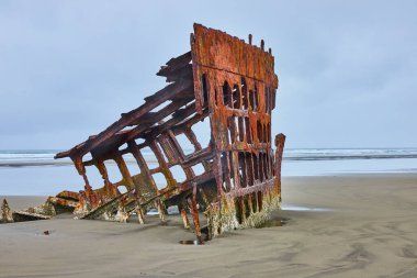 Hammond, Oregon 'da ıssız bir sahilde Peter Iredale' in enkazı. Paslı metal ve bulutlu gökyüzü, ABD 'nin batısındaki bu tarihi deniz kalıntısının unutulmaz güzelliğini ve izolasyonunu vurguluyor.