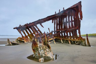 Hammond, Oregon sahilindeki Peter Iredale gemi enkazının paslı kalıntıları gri, bulutlu bir gökyüzünün altında. Çürümüş yapı ve sakin deniz, terk edilme, tarih ve doğanın ebedi güç temaları çağrıştırıyor..