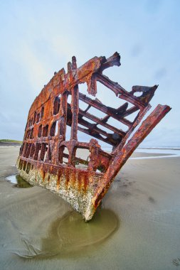 Oregon Hammond Sahili 'ndeki paslanmış Peter Iredale enkazının kasvetli sahnesi. Yüksek, aşınmış yapı bulutlu bir gökyüzüne karşı duruyor, Birleşik Devletler 'deki çürümeyi ve deniz tarihini simgeliyor.