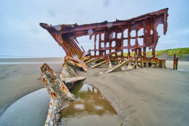 Hammond, Oregon 'daki engin kumlu bir sahilde Peter Iredale' in enkazından geriye kalanlar. Paslı kirişler ve paneller bulutlu bir gökyüzünün ortasında duruyor, tarihin, çürümenin ve doğanın geri kazanma temalarını anımsatıyor.