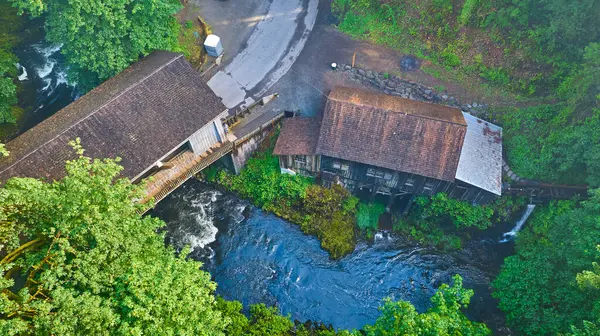 stock image Aerial view of Cedar Creek Grist Mill in Washington, showcasing rustic wooden buildings connected by a bridge over a scenic river. Surrounded by lush woodland, this historical site evokes nature and