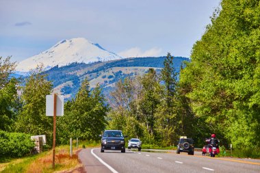 Berrak bir gökyüzünün altında arabaları ve motosikleti olan, yemyeşil ağaçlar ve Oregon 'daki görkemli kar kaplı Hood Dağı tarafından çerçevelenmiş manzaralı bir otoyol. Seyahat, macera ve doğa temalı projeler için mükemmel..