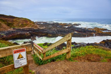 Rugged coastline of Thors Well, Oregon, where powerful waves crash against jagged rocks. A weathered wooden fence and warning sign highlight the need for caution in this dramatic natural setting. clipart