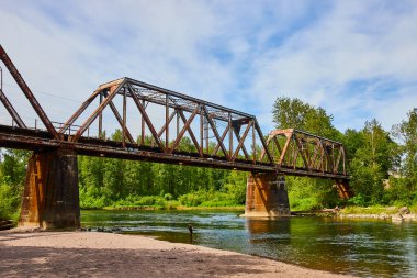 Troutdale, Oregon 'daki Sandy Nehri üzerindeki Rustik köprü. Yıpranmış metal yapısı açık mavi gökyüzünün altındaki yemyeşil ile çelişiyor. Yalnız bir birey dingin suda yürür.