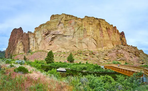 stock image Serene river and lush greenery frame the majestic rock formations of Smith Rock State Park in Oregon. A wooden footbridge invites exploration, blending human craftsmanship with natural beauty. Perfect