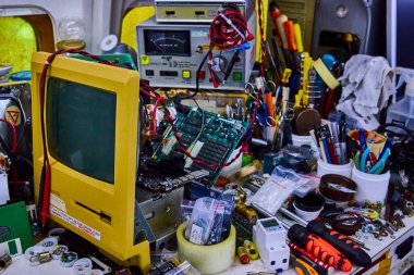 Antika ve modern teknolojiyle dolu hareketli bir elektronik tamir atölyesi. Sarı monitör ve dijital cihazlar tarafından vurgulanan bu alan, yenilikleri ve eski elektronikleri canlandırma sanatını somutlaştırıyor..