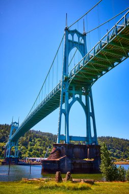 Portland, Oregon 'daki St. Johns Köprüsü' nde yemyeşil kuleler ve dingin bir su gövdesi üzerinde açık mavi gökyüzü altında Gotik tarzı mimari sergileniyor. Seyahat, mühendislik ve...