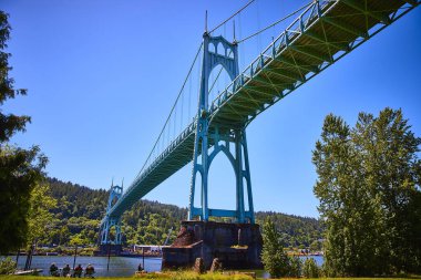 Portland 'daki St. Johns Köprüsü açık mavi gökyüzünün altındaki Willamette Nehri' ni kapsar. Gotik kemerler ve deniz mavisi kuleler göz kamaştırıcı bir manzara yaratır. Yakınlarda, insanlar güneşli bir günün tadını çıkarıyorlar, bir tutam topluluk ekliyorlar ve